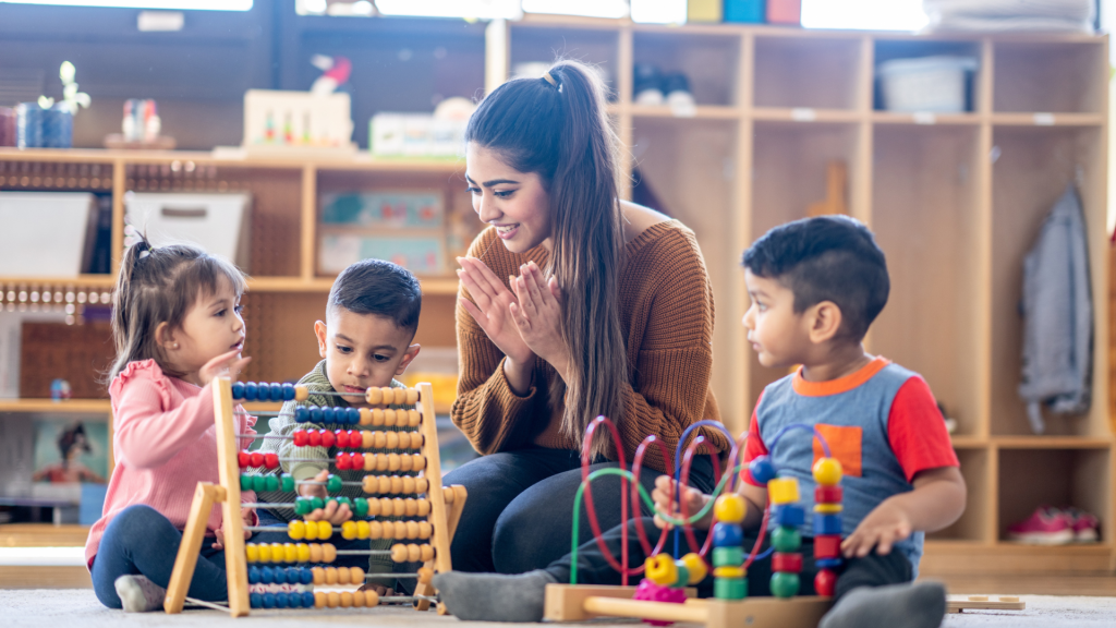A female Kindergarten teacher of Middle Eastern decent, sits on the floor with students as they play with various toys and engage in different activities. They are each dressed casually as they learn through their play.