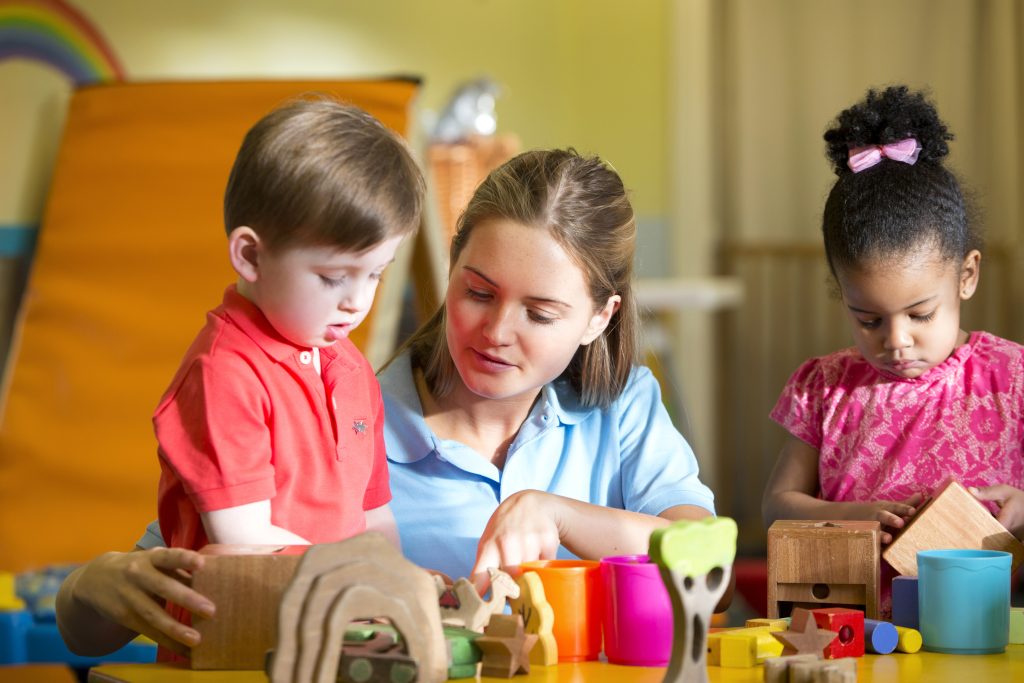 A young female teacher is supervising two children in the nursery playing with building blocks, the children are content playing. The sun is shining through the window and shadows can be seen on the faces of the teacher and children.